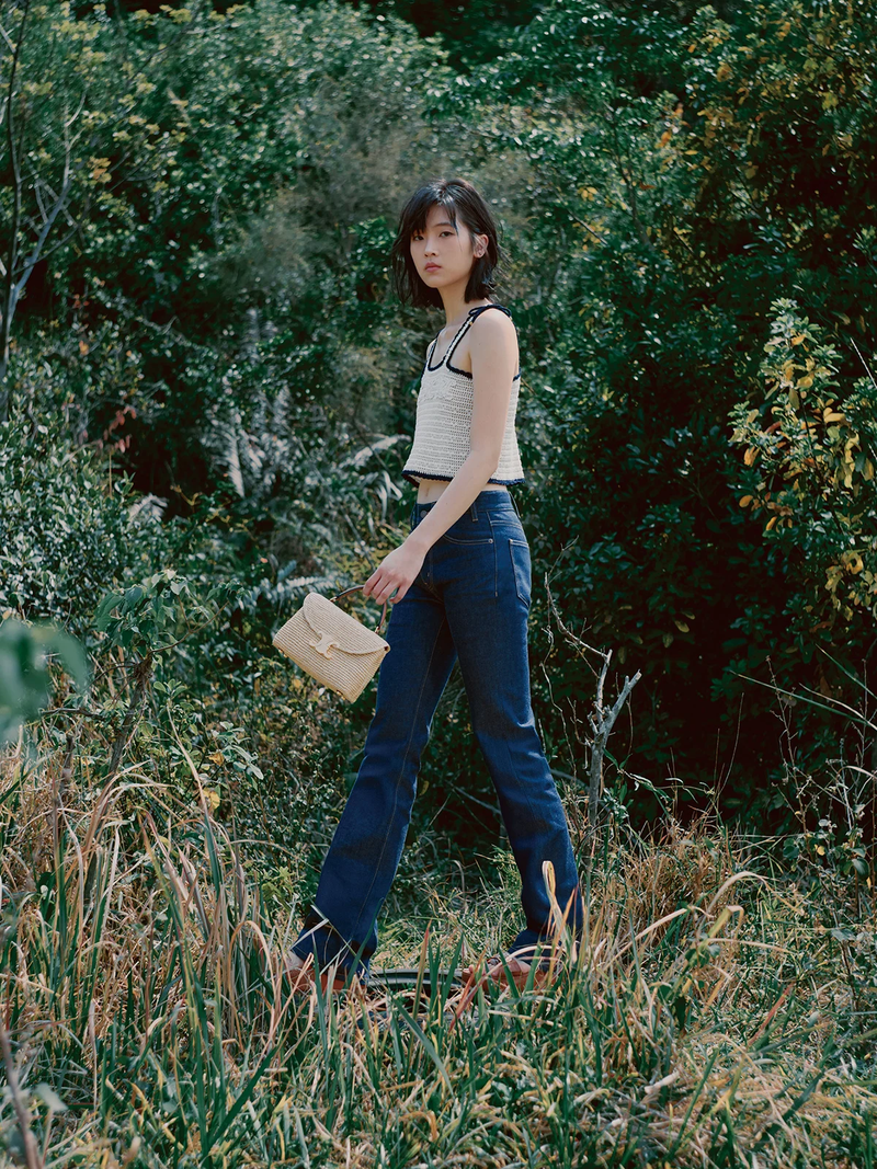 Young woman in a white knitted top and blue jeans walking through a grassy field with a beige purse and a background of dense green trees.