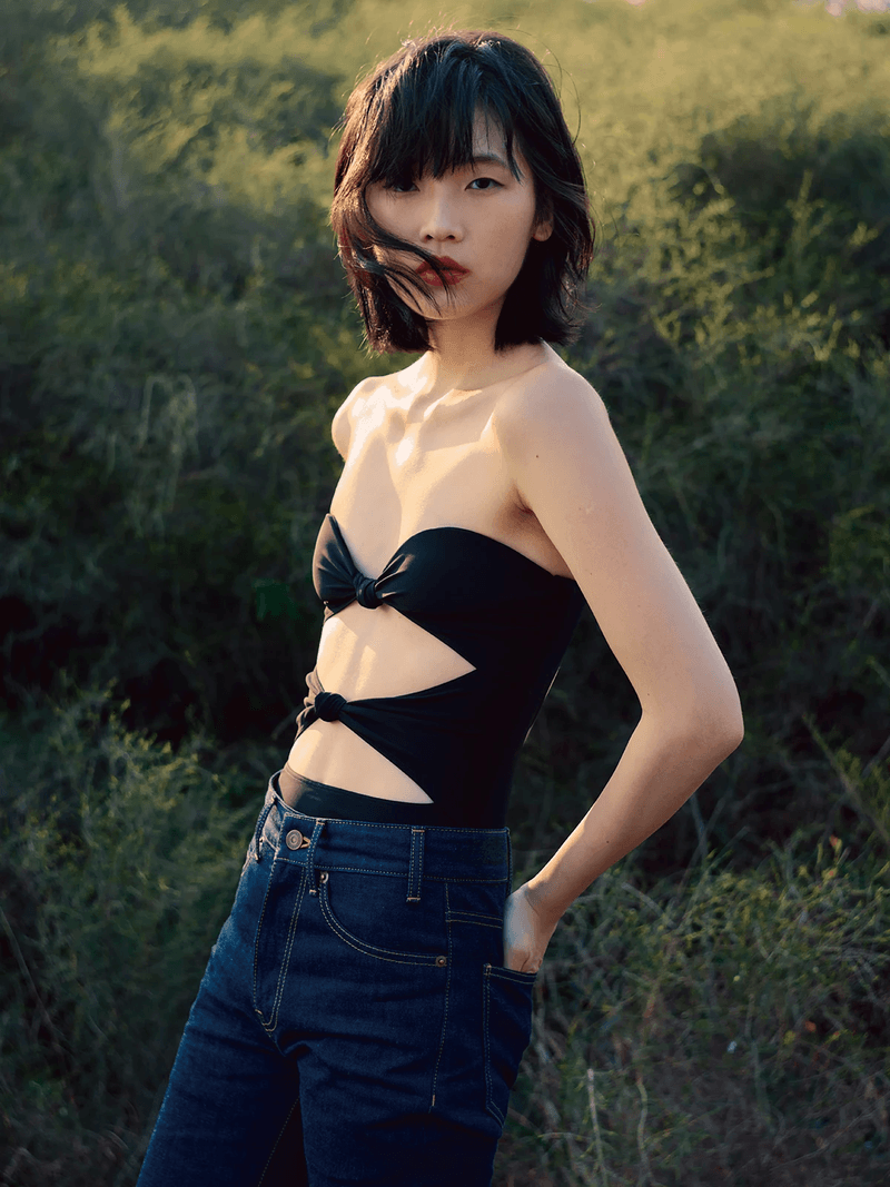 Young woman with dark hair poses in a black strapless cut-out top and blue jeans outdoors in natural lighting, looking at the camera.