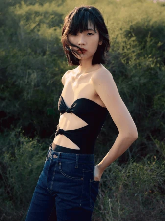 Young woman with dark hair poses in a black strapless cut-out top and blue jeans outdoors in natural lighting, looking at the camera.