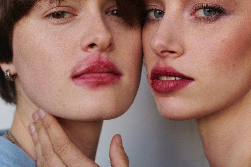 Close-up shot of two young women with pink lipstick on their lips, highlighting their facial features and smooth skin tones in soft lighting.