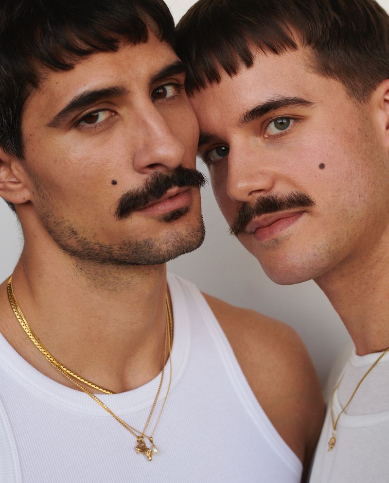 Close-up portrait of two adult men with moustaches, wearing tank tops and gold necklaces against a light neutral background.