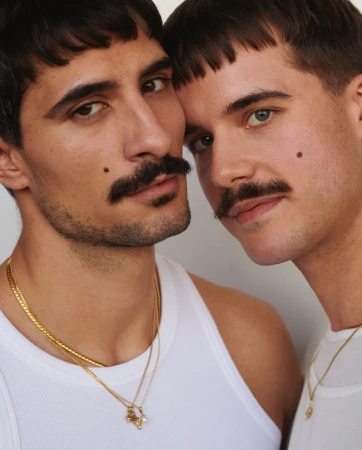Close-up portrait of two adult men with moustaches, wearing tank tops and gold necklaces against a light neutral background.