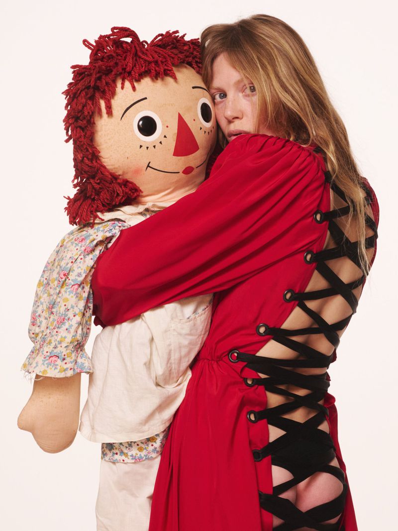 A young adult woman with long blonde hair embraces a Raggedy Ann doll, wearing a red dress with a crisscross black lace-up back, white backdrop.