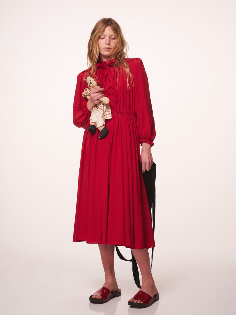 A full-length studio shot of a woman in a red dress holding a doll, complemented by black sandals and a black bag.