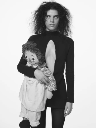 Young woman in a black dress poses in front of a white background, holding a Raggedy Ann doll. The black and white studio shot is in portrait.