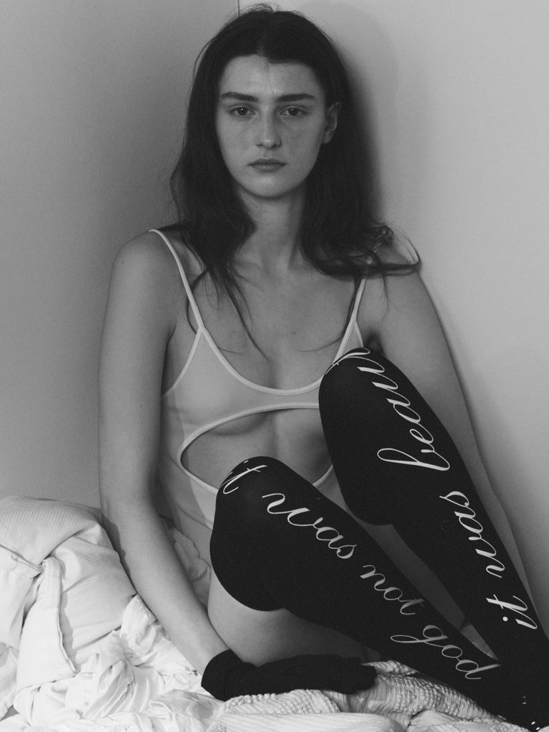 A young adult female with long brown hair poses indoors in a neutral-toned bodysuit and patterned thigh-high stockings, sitting on white bedding.