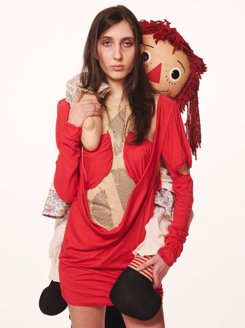 Brunette adult female with a neutral expression wearing a red dress and holding a Raggedy Ann doll, shot against a white backdrop.