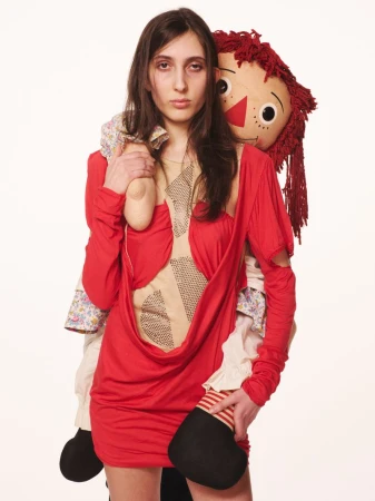 Brunette adult female with a neutral expression wearing a red dress and holding a Raggedy Ann doll, shot against a white backdrop.