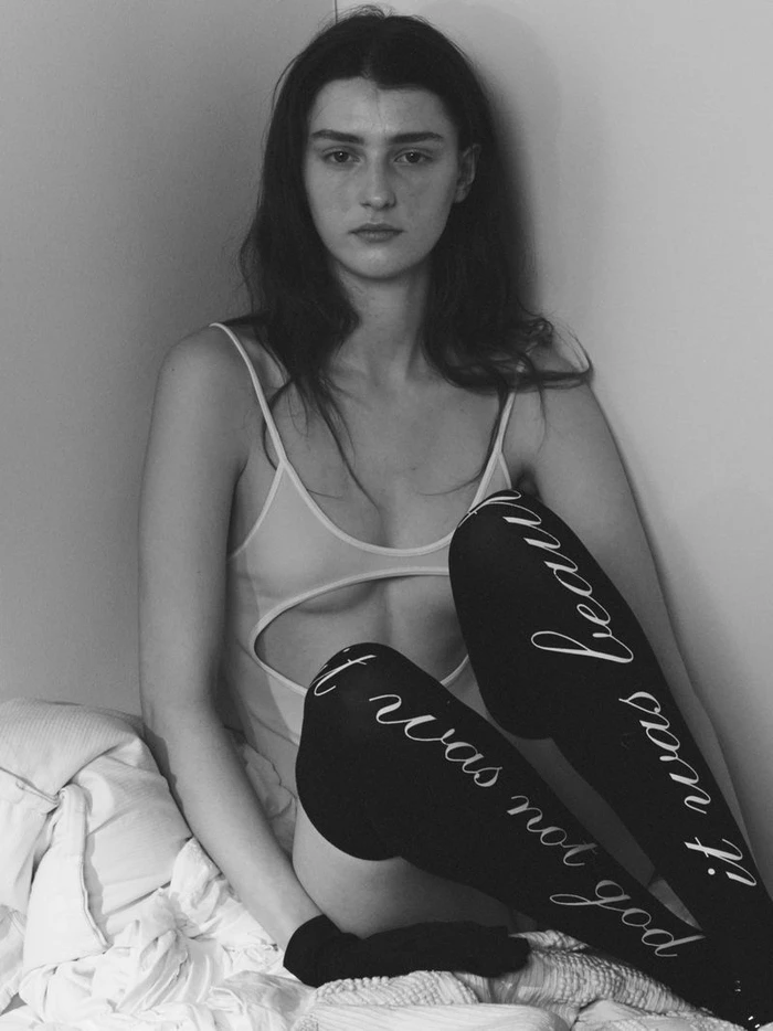 Monochrome portrait of a female model wearing printed thigh-high socks and a bodysuit seated against a white wall.