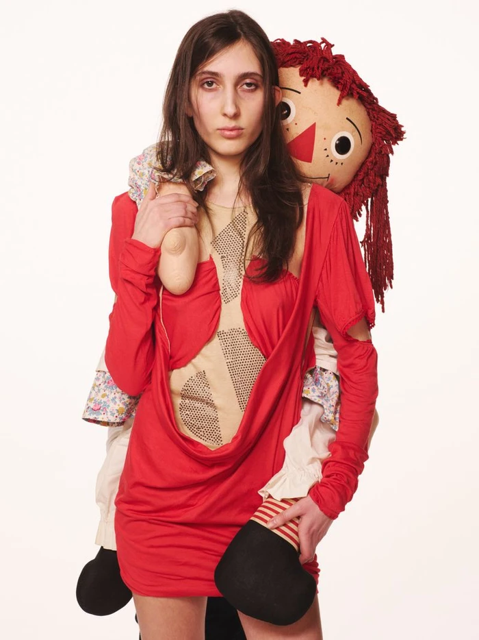 Model wearing a red dress carrying a large rag doll on her back against a plain white background