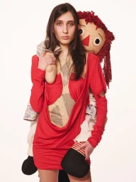 Model wearing a red dress carrying a large rag doll on her back against a plain white background