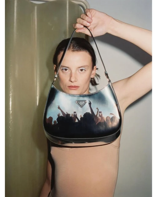 Woman holds a black Prada handbag featuring a crowd scene graphic, complemented by a sleek, beige bodysuit and minimalist makeup.
