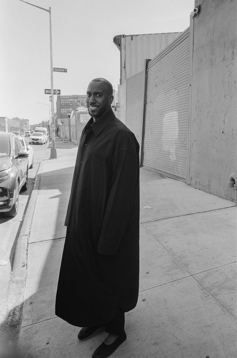 An African-American man in a long black coat stands on a city sidewalk on a sunny day, with buildings visible in the background.