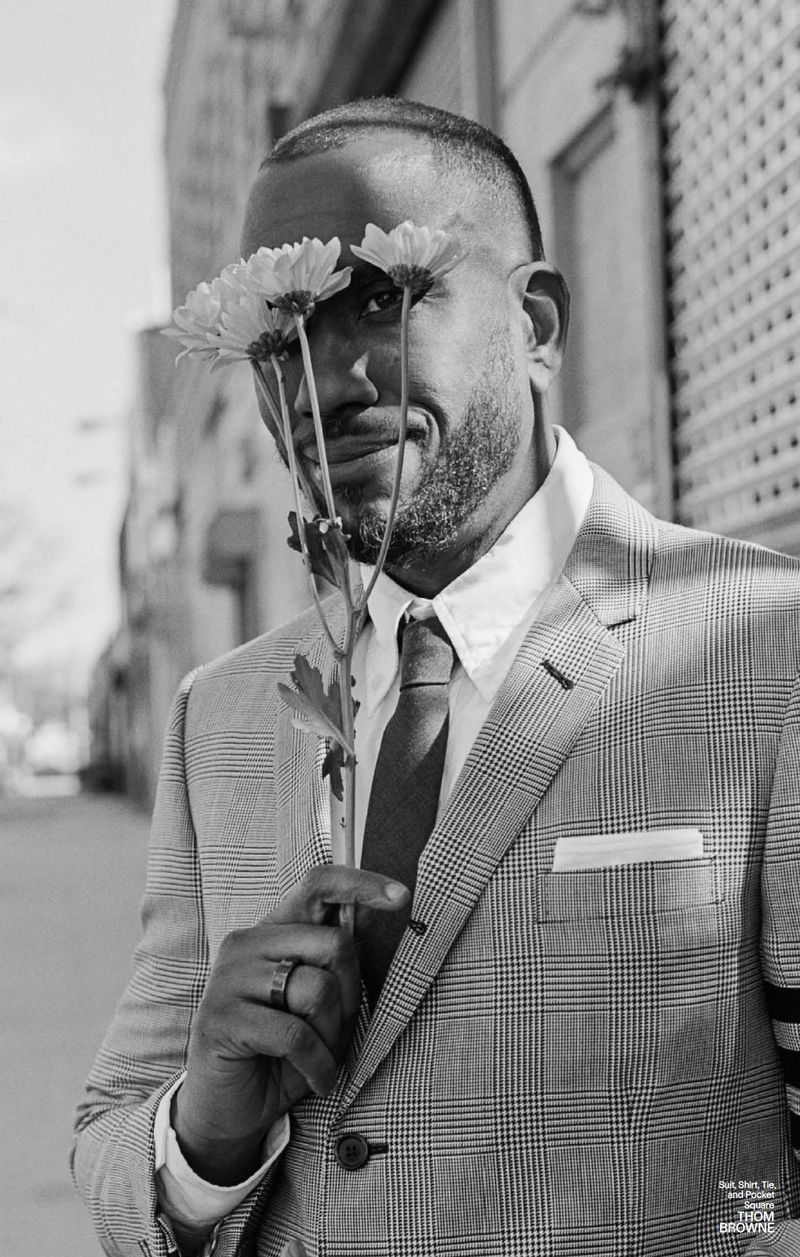 Black man in a grey plaid suit poses outside with daisies covering his eyes, creating a fashionable and playful black and white portrait.