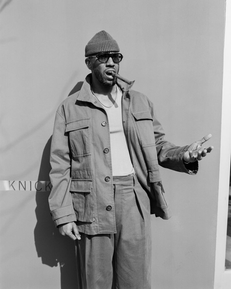 Monochrome portrait of adult male smoking a cigar, wearing a knit cap, sunglasses, jacket, pants, and necklace against a flat background.