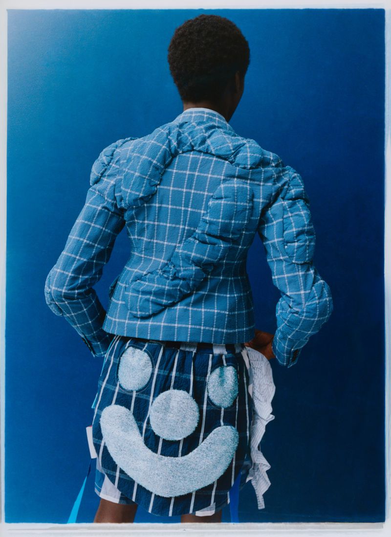 Woman in a blue and white checkered jacket and skirt with smiley face detail, standing in front of a royal blue backdrop.