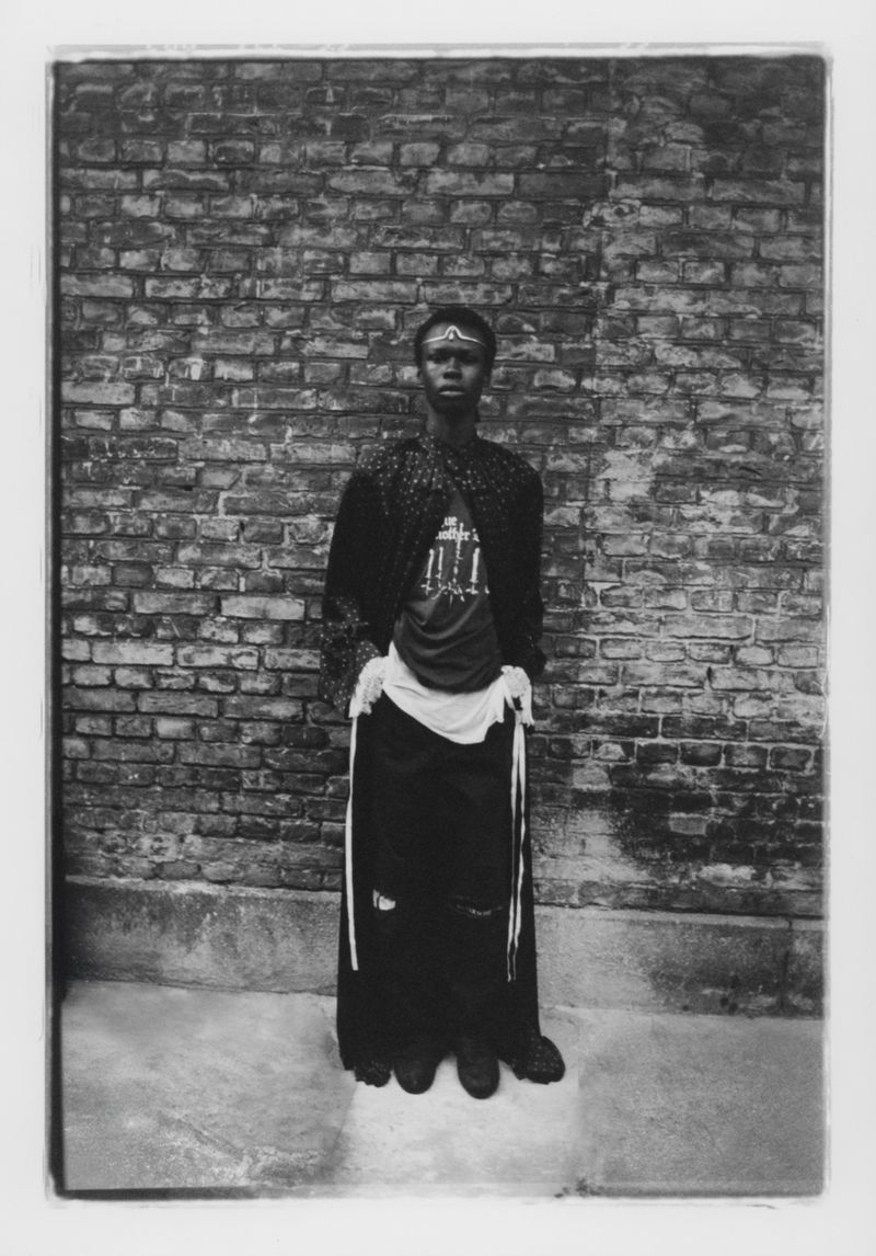 Monochrome portrait of a person with adorned forehead, patterned robe, and dark apparel standing before an old brick wall outdoors.