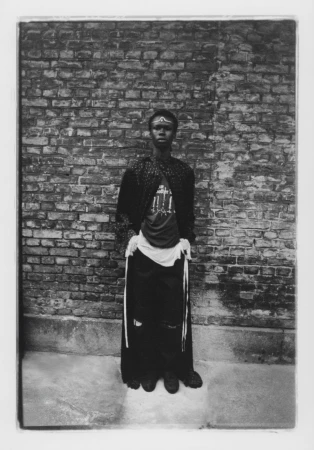 Monochrome portrait of a person with adorned forehead, patterned robe, and dark apparel standing before an old brick wall outdoors.