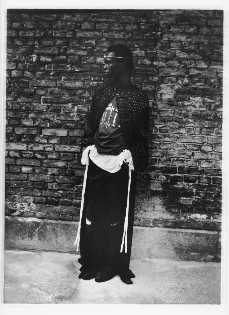 Monochrome portrait of a Black adult male in patterned jacket leaning against a brick wall, gazing left with neutral expression.