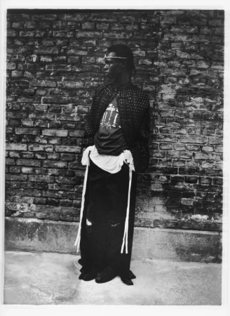 Monochrome portrait of a Black adult male in patterned jacket leaning against a brick wall, gazing left with neutral expression.