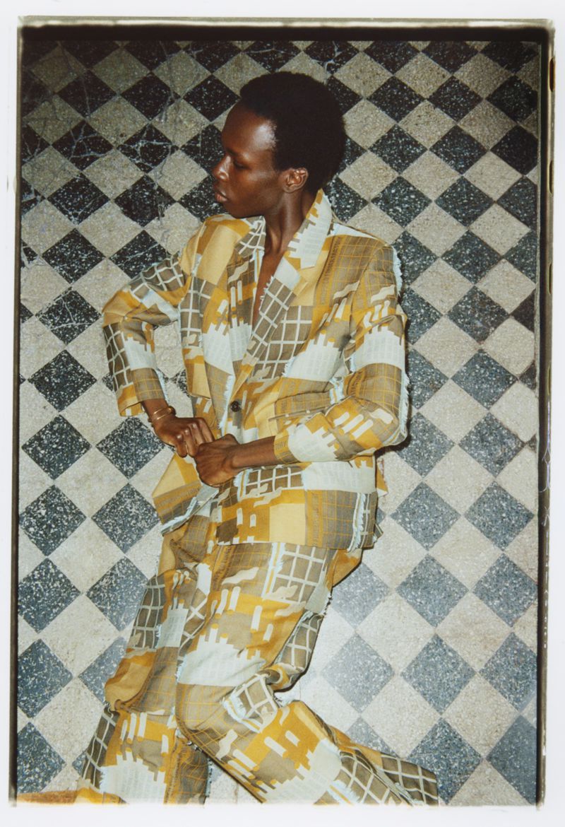 Fashionable adult male poses indoors wearing a tan and white patterned suit against a diamond-patterned tile background.