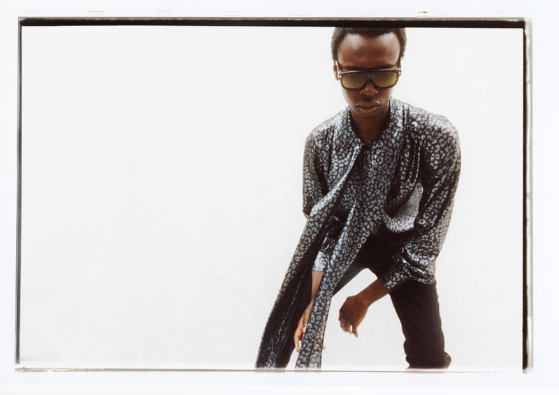 A person wearing sunglasses and a patterned shirt poses in front of a white backdrop, looking down, with a cool and fashionable aesthetic.