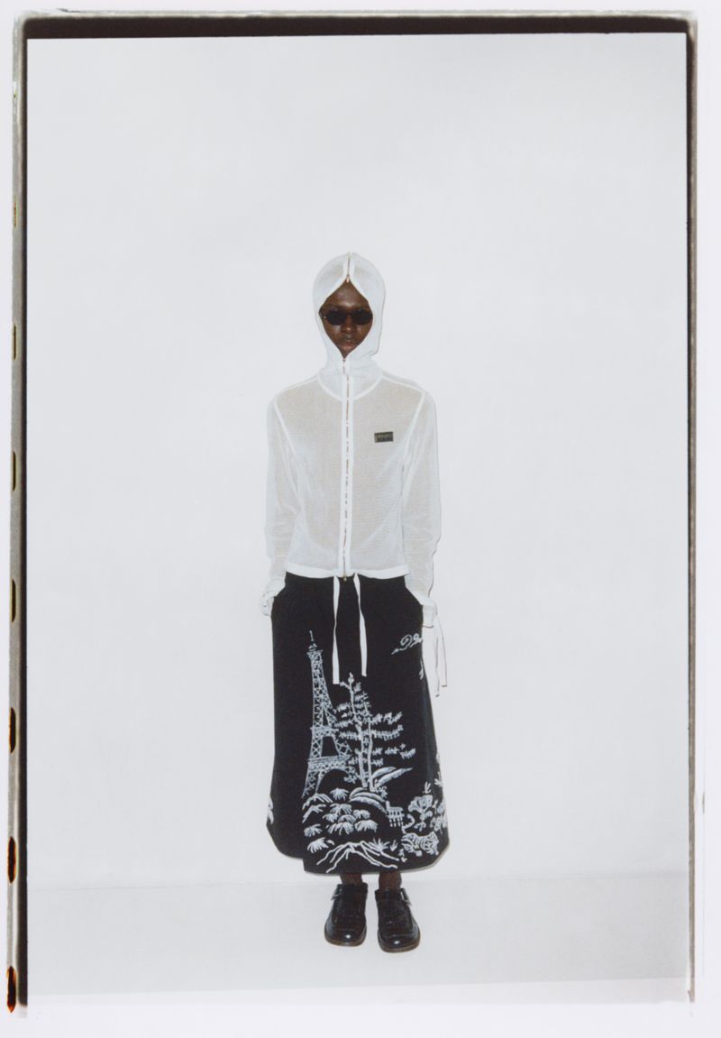 Fashion shot of a model wearing a white hooded jacket and black skirt featuring an Eiffel Tower graphic against a bright white backdrop.