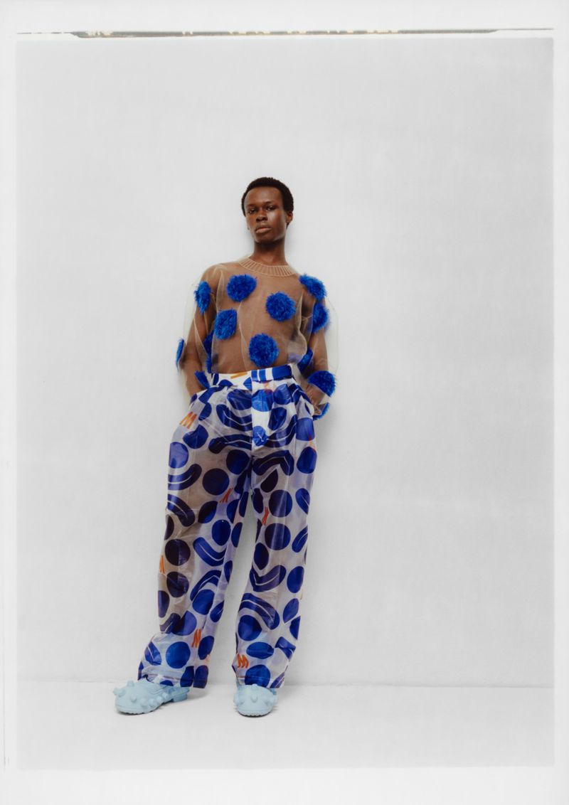 A slender black man in patterned blue and tan clothing strikes a pose against a white wall, showcasing a unique fashion-forward style.