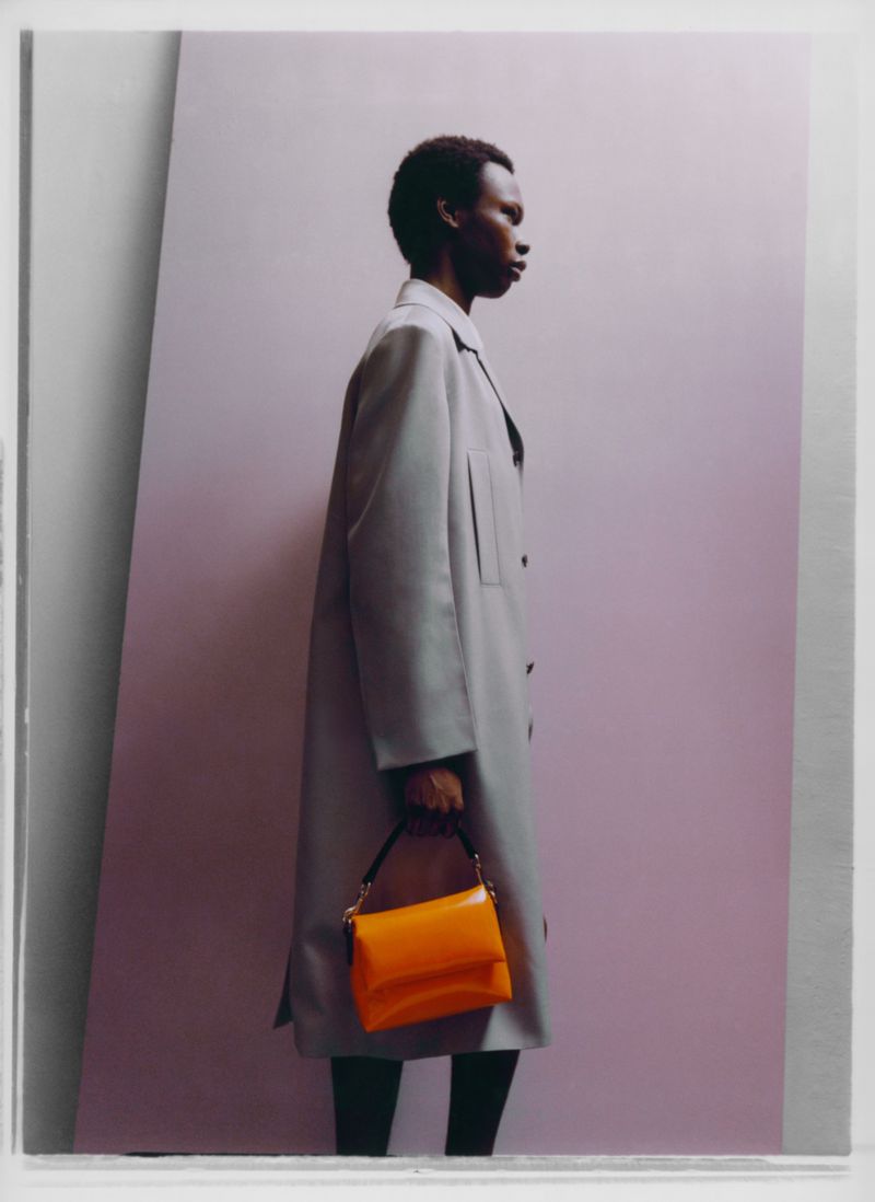 Fashion editorial featuring a Black male model in a grey coat and orange handbag, set against a purple backdrop with geometric shadows.