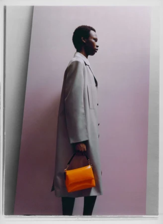 Fashion editorial featuring a Black male model in a grey coat and orange handbag, set against a purple backdrop with geometric shadows.