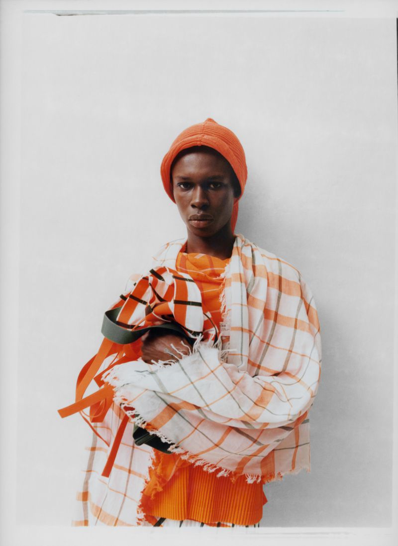 Fashion model poses in orange knit hat, top, and white plaid scarf against a white wall. The composition is a medium shot.