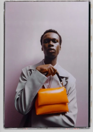 A person models an orange handbag. The individual wears a grey blazer, and the background is a light purple. The image focuses on fashion.
