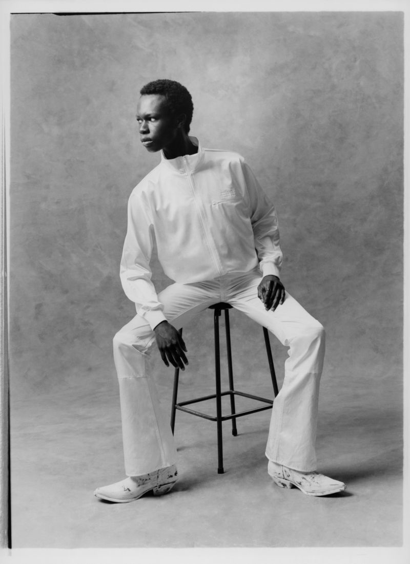 Black and white portrait of African American male fashion model sitting on a stool in studio wearing light colored shirt and pants.