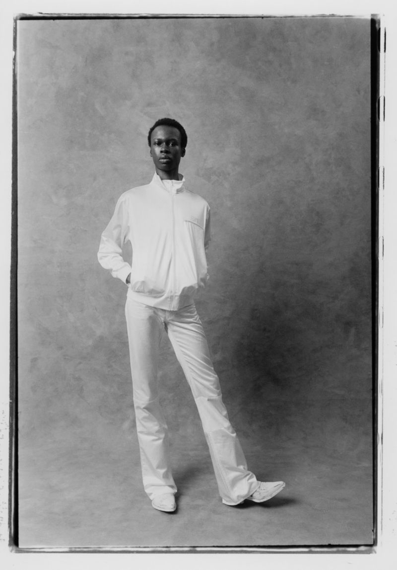 A young African-American man in a white tracksuit poses against a mottled gray background in a studio shot, full body, casual style.