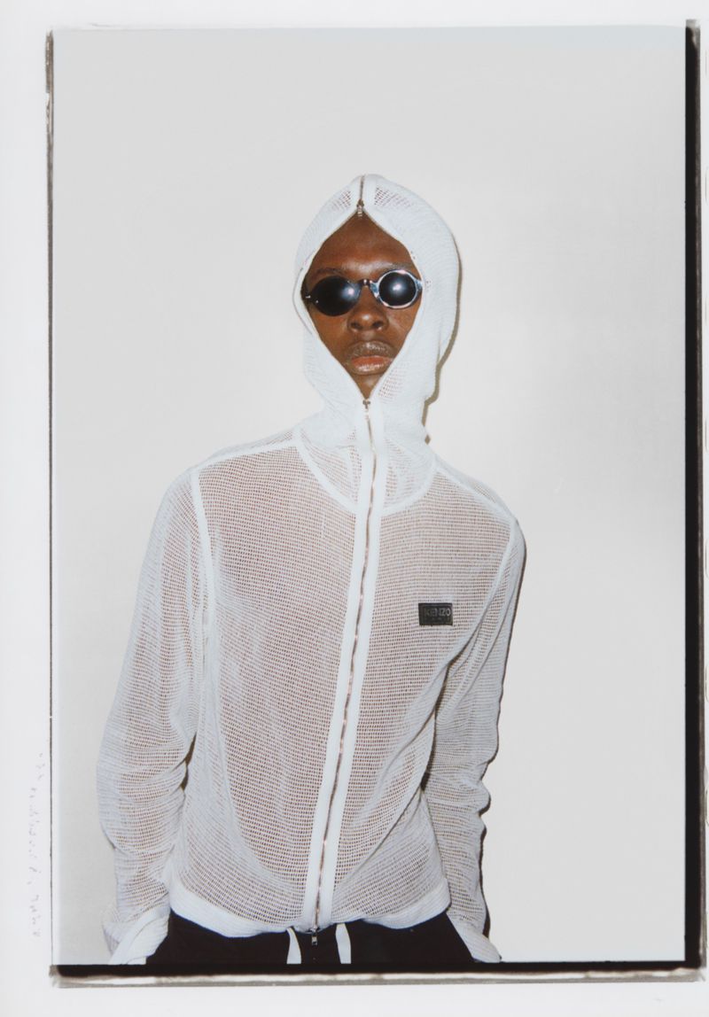Stylish adult male in sunglasses and a white hooded mesh jacket poses indoors against a white wall, fashion portrait.