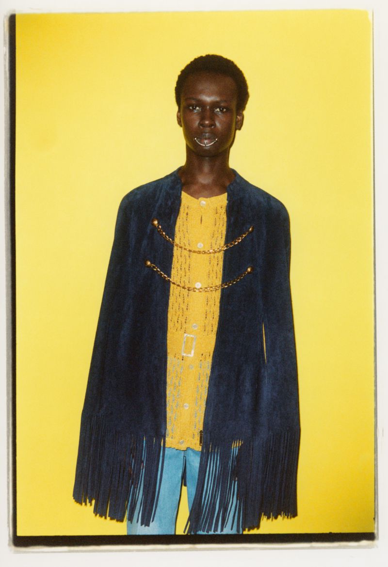 Adult male model in a yellow button-down shirt, blue pants, and blue fringe jacket posing against a yellow background in a studio.