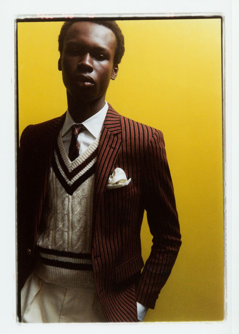 Portrait of a Black male model wearing a striped blazer, sweater vest, tie, and white dress pants against a yellow background.
