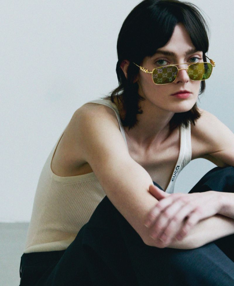 Portrait of a young adult woman wearing sunglasses with a Gucci logo pattern, a tank top, and dark pants against a neutral backdrop.