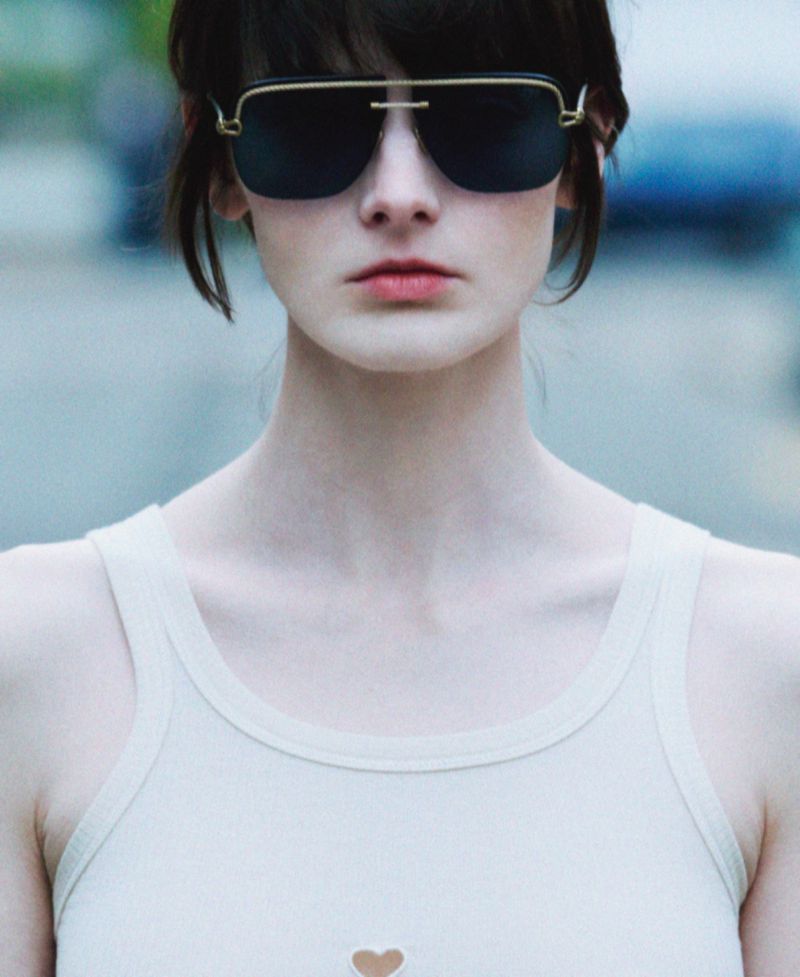 Close-up of an adult woman wearing sunglasses and a white tank top, with a blurred background. She has dark hair and fair skin.