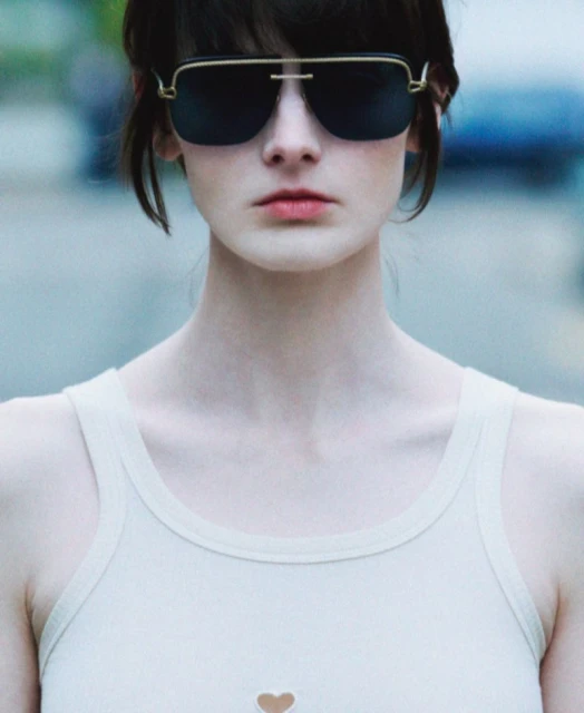 Close-up of an adult woman wearing sunglasses and a white tank top, with a blurred background. She has dark hair and fair skin.
