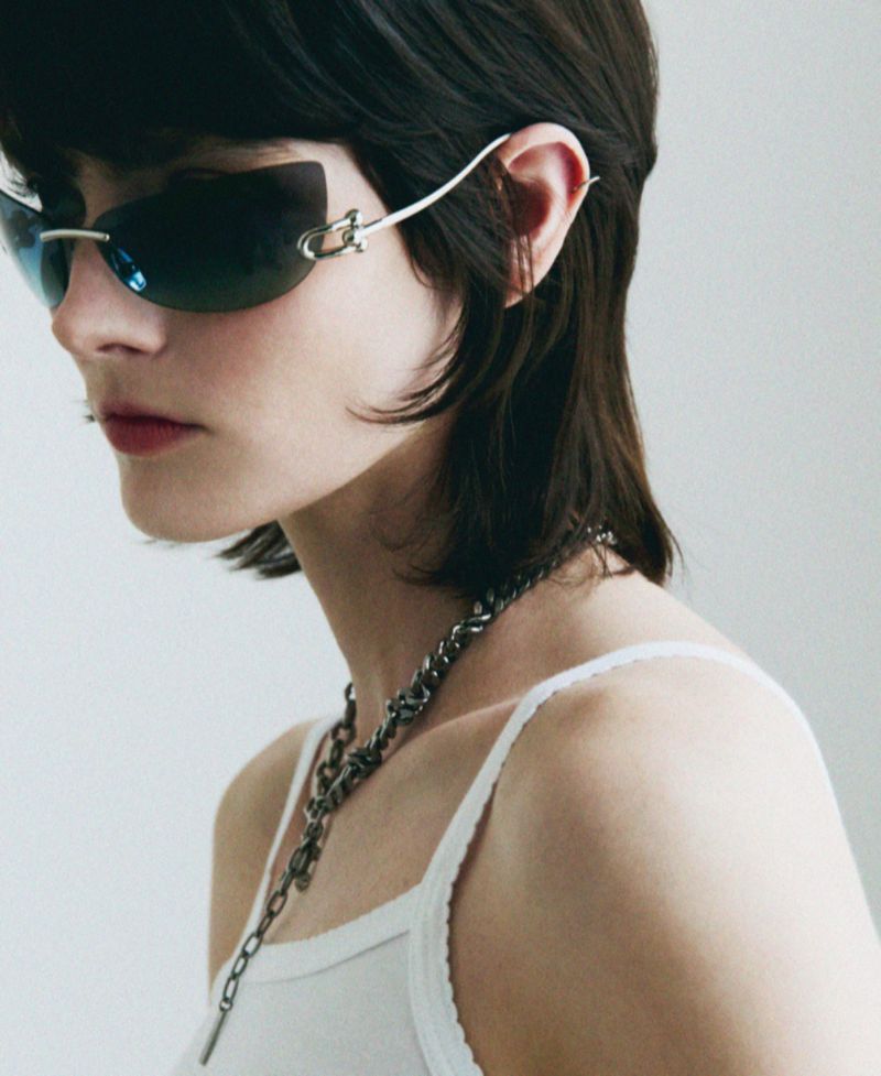 Close-up studio portrait of a young woman with dark hair and sunglasses, wearing a white tank top and a silver necklace.
