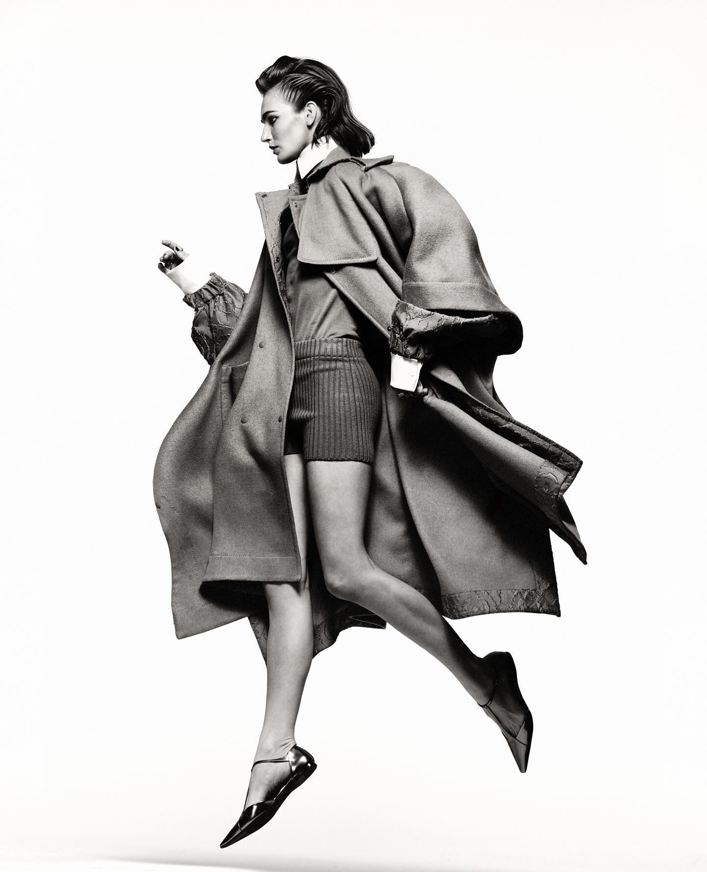 Fashion editorial shot in black and white featuring a woman jumping mid-air wearing a long coat and flats against a white backdrop.
