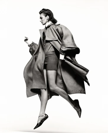 Fashion editorial shot in black and white featuring a woman jumping mid-air wearing a long coat and flats against a white backdrop.