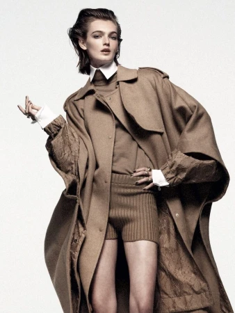 A fashion portrait showcases an adult woman in a brown trench coat, sweater, and knit shorts against a clean white backdrop, emphasizing modern style.