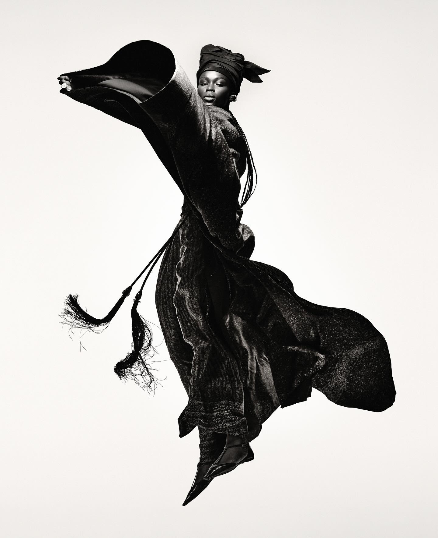 Fashion shot of a Black female model in draped black fabric, wearing a turban and ballet flats, posed against a stark white backdrop.