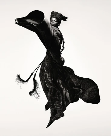 Fashion shot of a Black female model in draped black fabric, wearing a turban and ballet flats, posed against a stark white backdrop.