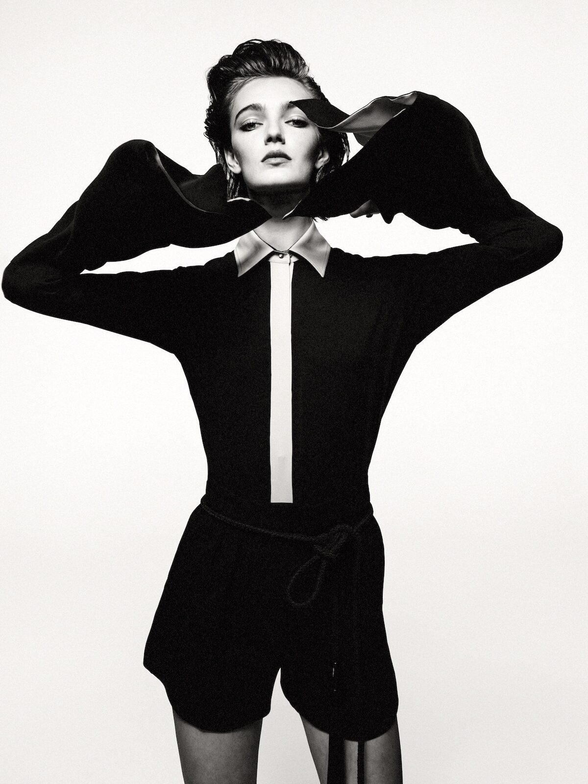 Monochrome studio portrait of a young adult woman in a black romper with dramatic sleeves, standing against a stark white backdrop.