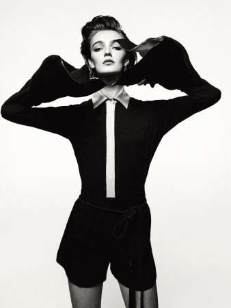 Monochrome studio portrait of a young adult woman in a black romper with dramatic sleeves, standing against a stark white backdrop.