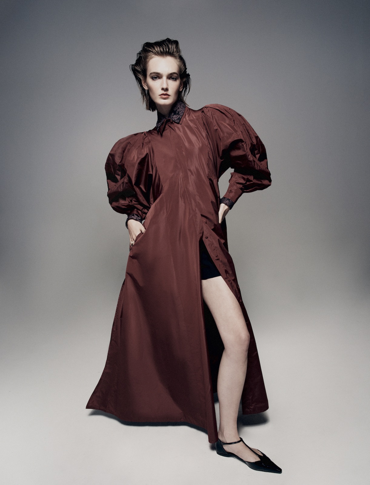 Elegant female fashion model posing in a maroon dress with puffy sleeves and black flats against a grey backdrop in a photo studio.