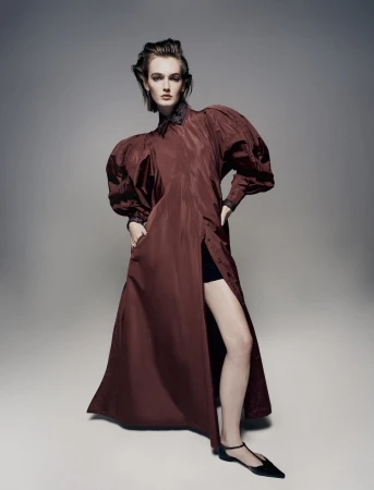 Elegant female fashion model posing in a maroon dress with puffy sleeves and black flats against a grey backdrop in a photo studio.